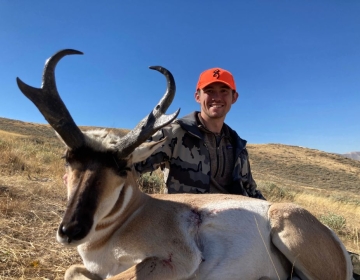 A hunter wearing a detailed camo jacket proudly showcases a trophy antelope, set against brilliant blue sky, completing another memorable SNS-guided adventure.